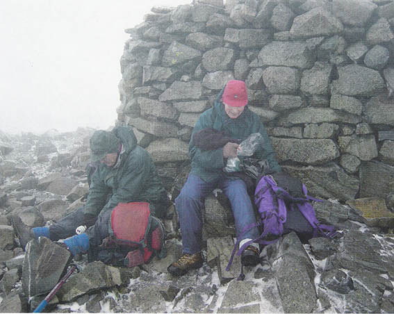 John and Allan take a lunch break in the snow (at what looks like the summit cairn on Scafell Pike)
